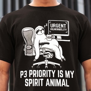 Op de afbeelding: Zwart t-shirt met een witte illustratie van een luiaard in een bureaustoel, met de tekst "URGENT VULNERABILITY" op een computerscherm. De tekst "P3 PRIORITY IS MY SPIRIT ANIMAL" staat eronder.