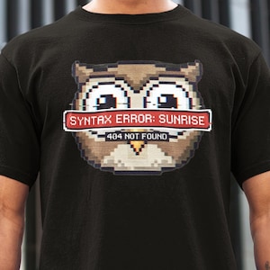 Könnte beinhalten: Schwarzes T-Shirt mit einem pixeligen Eulen-Design. Der Aufdruck zeigt den Text "SYNTAX ERROR: SUNRISE" auf einem roten Banner und "404 NOT FOUND" darunter. Die Eule ist braun, beige und weiß.
