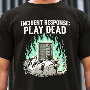 Op de afbeelding: Zwart t-shirt met de witte tekst "INCIDENT RESPONSE: PLAY DEAD". Een cartoon opossum met zonnebril ligt op zijn rug voor een serverrack omringd door groene vlammen. De opossum is grijs en wit.