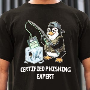 May include: Black t-shirt featuring a cartoon penguin wearing a cap and vest, holding a fishing rod with a floppy disk labeled "ADMIN PASSWORD" as bait. The text "CERTIFIED PHISHING EXPERT" is printed below the graphic.