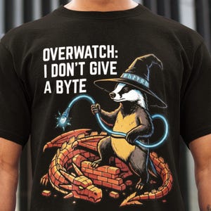 May include: Black t-shirt with the text "OVERWATCH: I DON'T GIVE A BYTE". The graphic features a badger wearing a wizard hat, holding a glowing blue cable, and sitting on a brick structure with a dragon.