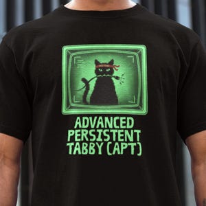 Puede incluir: Camiseta negra con un gato negro pixelado con una cinta roja en la cabeza y un cable verde brillante, dentro de una pantalla verde. El texto "ADVANCED PERSISTENT TABBY (APT)" se muestra debajo del gráfico.