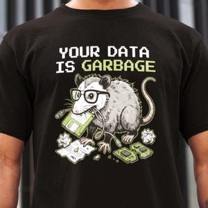 Może przedstawiać: Czarny t-shirt z napisem "YOUR DATA IS GARBAGE" w białej i zielonej pikselowej czcionce. Centralnym elementem graficznym jest rysunkowy opos z okularami i dyskietką, otoczony komponentami komputerowymi.