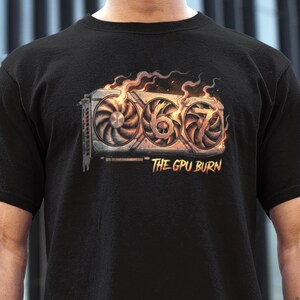 May include: Black t-shirt featuring a graphic of a computer graphics card engulfed in flames. The design includes the numbers "667" and the text "THE GPU BURN" in a stylized font. The shirt is made of a soft material.