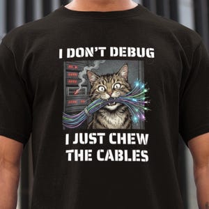 Può includere: T-shirt nera con una grafica di un gatto tigrato che tiene in bocca un fascio di cavi colorati. Il testo "I DON'T DEBUG I JUST CHEW THE CABLES" è stampato sopra e sotto l'illustrazione del gatto.