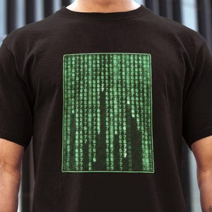 Op de afbeelding: Zwart T-shirt met een groen digitaal regenontwerp, dat doet denken aan een computercode matrix. De afbeelding is een rechthoekig raster van groene getallen en symbolen. Het shirt heeft een klassieke ronde hals.