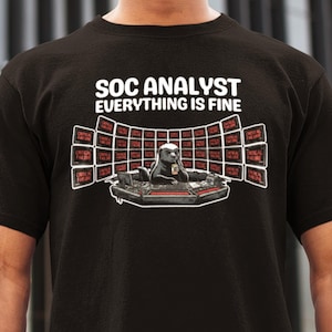Op de afbeelding: Zwart t-shirt met de witte tekst "SOC ANALYST EVERYTHING IS FINE". De afbeelding toont een das in een controlekamer met schermen met "CRITICAL FAILURE". De das houdt een drankje vast.
