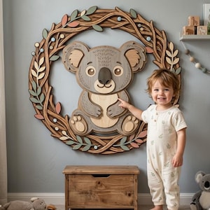 May include: A wooden koala bear wall art piece, surrounded by a wreath of leaves and branches. The koala is gray and tan, with a friendly expression. The wreath features green, pink, and blue accents. A child points at the koala.