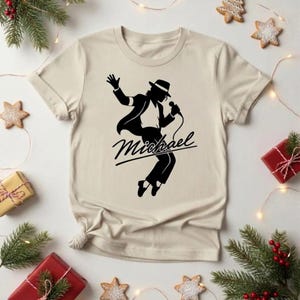 May include: A beige short-sleeved t-shirt featuring a black graphic print of a person in a fedora hat and suit dancing while holding a microphone. The word Michael is printed in a cursive script font across the center of the graphic design.