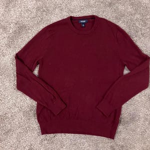 May include: A burgundy Lands' End crew neck sweater. The long-sleeved sweater is made of a soft material and has a ribbed hem and cuffs. The sweater is a solid colour and is displayed on a neutral-coloured surface.