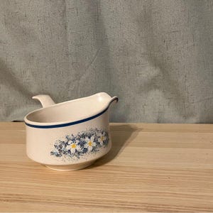 Op de afbeelding: Crèmekleurige keramische juskom met een blauwe streep en bloemmotief. De kom heeft een handvat en een tuit. Het bloemmotief heeft blauwe en witte bloemen met gele centra. De juskom staat op een houten oppervlak.