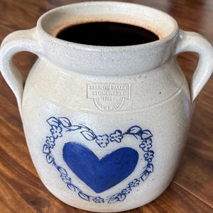 May include: A beige stoneware crock with two side handles and a dark interior. The front features a blue painted heart surrounded by a floral vine pattern. A stamped mark reads SALMON FALLS STONEWARE 1992 DOVER NH in the center of the vessel.