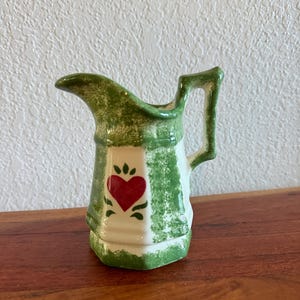 Puede incluir: Una pequeña jarra de cerámica con un diseño a rayas verdes y blancas. Un corazón rojo está centrado en la parte delantera, con detalles en verde. El asa y el borde también son verdes. La jarra está sobre una superficie de madera.