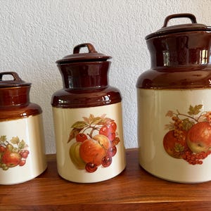 Pode incluir: Conjunto de três potes de cerâmica vintage com tampas. Os potes são castanhos e creme, com um desenho de frutas. O pote maior tem aproximadamente 25 cm de altura, o médio 20 cm e o menor 15 cm.