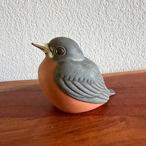 May include: A ceramic bird figurine with a gray head and wings, a peach-colored body, and a gold-colored beak. The bird is perched on a wooden surface.