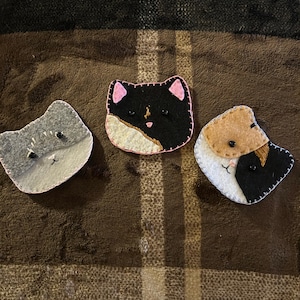 Op de afbeelding: Drie handgemaakte katvormige broches van vilt. Eén is grijs, één is zwart-wit en de andere is bruin, zwart en wit. Elke broche heeft geborduurde details en een gestikte omtrek. De broches worden gepresenteerd op een bruin, getextureerd oppervlak.