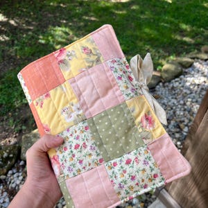 Puede incluir: Una funda de diario acolchada hecha a mano con un diseño de patchwork. La funda presenta cuadrados de tela melocotón, amarilla, rosa y verde con estampados florales y de lunares. Una cinta color crema está atada alrededor del diario.