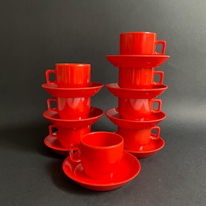 May include: A collection of eight vibrant red coffee cups and saucers. The cups feature square handles and are arranged in stacks of two and three. The saucers are circular and gently curved. The backdrop is a plain black.