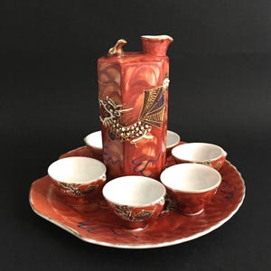 Può includere: Un set da sake giapponese rosso e oro. Il set comprende una caraffa rettangolare con un motivo a drago, sei piccole tazze e un vassoio rotondo. Le tazze e il vassoio hanno una combinazione di colori rosso e oro.