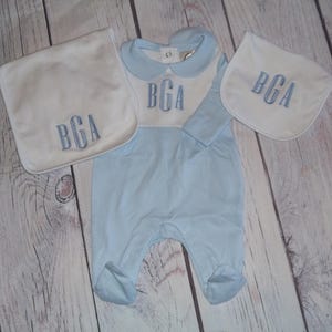 May include: A baby clothing set in light blue and white, featuring a footed onesie and two bibs. Each item is embroidered with the initials "BGA" in a matching blue thread. The set is displayed on a light wood surface.