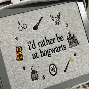 May include: A heather gray sweatshirt featuring black embroidered text that reads "i'd rather be at hogwarts" surrounded by small icons including glasses, a broomstick, an owl, a castle, a scarf, a cauldron, a 9 3/4 sign, and a wand.