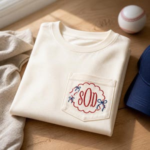 Op de afbeelding: Crèmekleurig T-shirt met een zakje met een honkbalthema. De zak heeft een rode geschulpte rand met geborduurde honkballen en strikken. De initialen "SOD" zijn in rood script geborduurd in de geschulpte rand. Een honkbal en een marineblauwe baseballpet zijn ook zichtbaar.
