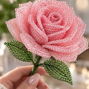 Könnte beinhalten: Eine handgefertigte Rosenskulptur aus kleinen, durchscheinenden rosa Glasperlen, die die Blütenblätter bilden, und schimmernden metallisch grünen Glasperlen für die Blätter, befestigt an einem mit grünem Material umwickelten Stiel.