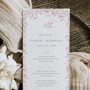 May include: A wedding menu card with a floral border and the names "FLORIA + NICHOLAS" and the date "August 25, 20XX". The menu lists a goat cheese salad, herb roasted chicken, slow roast pork loin, poached salmon, and cheesecake. The card is on a wooden charger with a white flower and draped fabric.