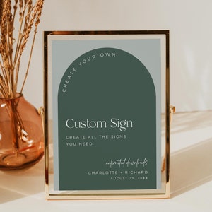 May include: A framed sign with a gold-colored metal frame. The sign has a sage green and light blue background with the text "Custom Sign" and "Create all the signs you need". The sign also includes the text "unlimited downloads" and names and a date.