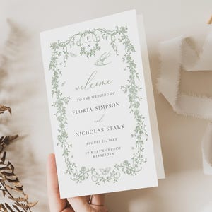 May include: A folded wedding invitation with a floral design in green. The invitation reads "welcome" and lists the names of the couple, the wedding date, and the church location. The invitation is held in a hand.