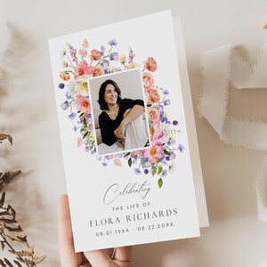 May include: A white memorial card featuring a photograph of a woman framed by a watercolour floral design in pinks, purples, and yellows. The card reads "Celebrating the Life of Flora Richards" with dates. The card is held by a hand.