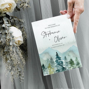 Op de afbeelding: Huwelijksuitnodiging met een aquarel-illustratie van een bergketen en dennenbomen. De tekst luidt "Welkom op de bruiloft van Stephanie + Oliver, 26 september 2021, St. Mary's Church, MN".