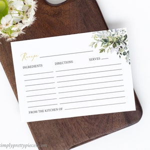 May include: A white recipe card with gold script that reads "Recipe", "Ingredients", "Directions", and "Serves". The card has a floral design in the upper right corner and is on a dark wooden cutting board. The text "FROM THE KITCHEN OF" is at the bottom.