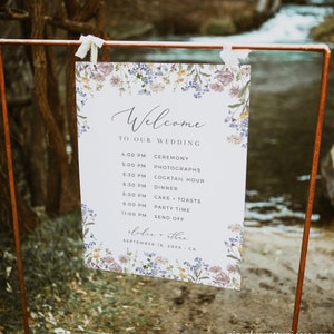 May include: A white wedding sign with a floral border and a list of events for the wedding day. The sign says "Welcome to our wedding" and lists the ceremony, photographs, cocktail hour, dinner, cake and toasts, party time, and send off. The sign also includes the names "Elodie + Ethan" and the date "September 16, 20XX - CA".