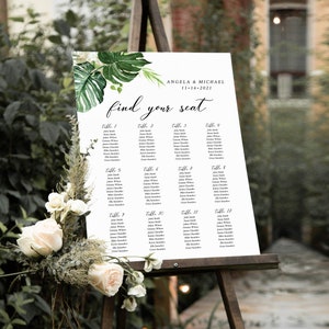 May include: A white wedding seating chart with green tropical leaves and the text "Angela & Michael 11-16-2021" and "find your seat". The chart lists guest names by table number.