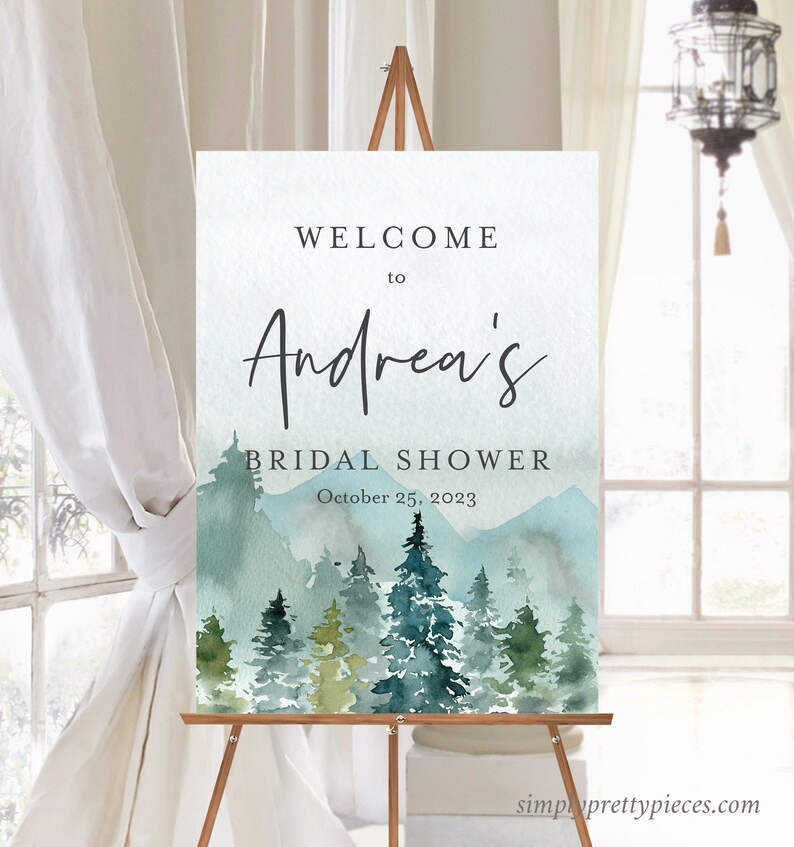 Op de afbeelding: Welkom bij Andrea's Bridal Shower bord met een aquarel-illustratie van een bergketen en dennenbomen in tinten blauw en groen. De tekst "October 25, 2023" staat onder de naam.