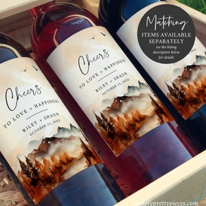May include: Two wine bottles with personalized labels featuring a watercolor mountain scene with a foggy lake. The labels say "Cheers to love and happiness" with the names Riley and Shaun and the date October 15, 20XX. The bottles are in a wooden crate.