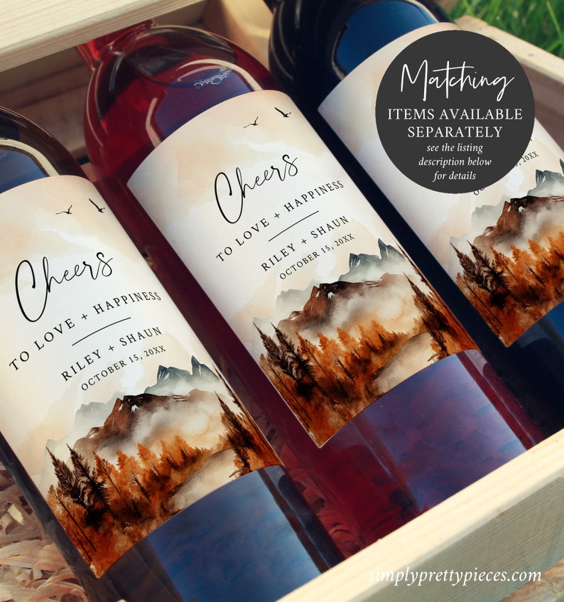 May include: Two wine bottles with personalized labels featuring a watercolor mountain scene with a foggy lake. The labels say "Cheers to love and happiness" with the names Riley and Shaun and the date October 15, 20XX. The bottles are in a wooden crate.
