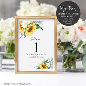 May include: A gold-framed table number card with a sunflower and white rose floral design. The card reads "table no. 1" and "Sophie & Michael August 25, 2022".