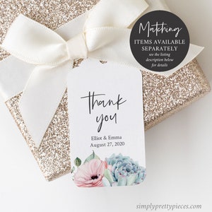 May include: A white thank you tag with a watercolour floral design featuring pink flowers and green succulents. The tag says "Thank you" in black script and "Elliot & Emma August 27, 2020". The tag is tied to a white satin ribbon on a gold glitter gift box.
