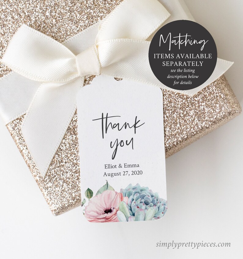 May include: A white thank you tag with a watercolour floral design featuring pink flowers and green succulents. The tag says "Thank you" in black script and "Elliot & Emma August 27, 2020". The tag is tied to a white satin ribbon on a gold glitter gift box.