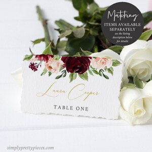 May include: A white place card with a floral design in burgundy, blush, and green. The card features the name "Laura Cooper" in gold script and "TABLE ONE" in a serif font. The card has a torn edge and is surrounded by white roses.