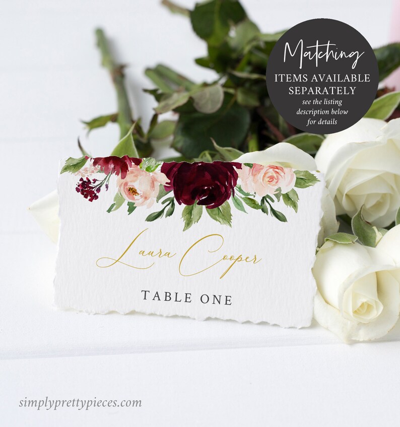 May include: A white place card with a floral design in burgundy, blush, and green. The card features the name "Laura Cooper" in gold script and "TABLE ONE" in a serif font. The card has a torn edge and is surrounded by white roses.