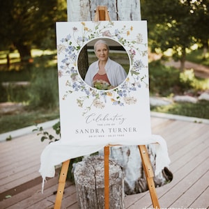 May include: A white sign with a floral wreath border and a photo of a woman in the centre. The sign reads "Celebrating the life of Sandra Turner 09.01.19XX - 08.22.20XX".