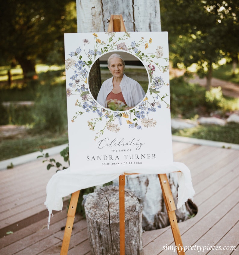 May include: A white sign with a floral wreath border and a photo of a woman in the centre. The sign reads "Celebrating the life of Sandra Turner 09.01.19XX - 08.22.20XX".