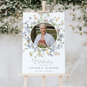 May include: A white memorial sign with a floral wreath border and a photo of a woman in the centre. The text reads "Celebrating the life of Sandra Turner 09.01.19XX - 08.22.20XX".