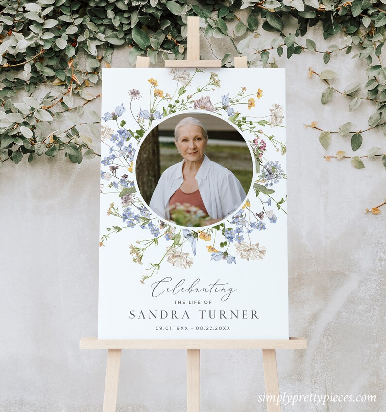 May include: A white memorial sign with a floral wreath border and a photo of a woman in the centre. The text reads "Celebrating the life of Sandra Turner 09.01.19XX - 08.22.20XX".