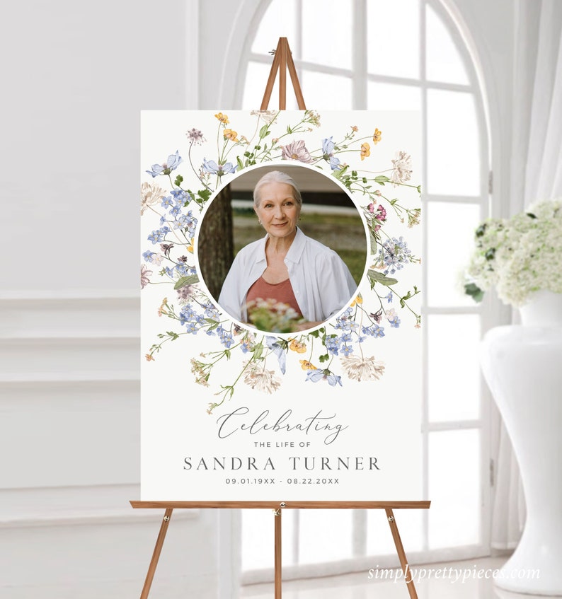 May include: A white memorial sign with a floral wreath design featuring blue and yellow flowers. A photo of a woman is in the centre of the wreath. The text "Celebrating the life of Sandra Turner 09.01.19XX - 08.22.20XX" is at the bottom of the sign.