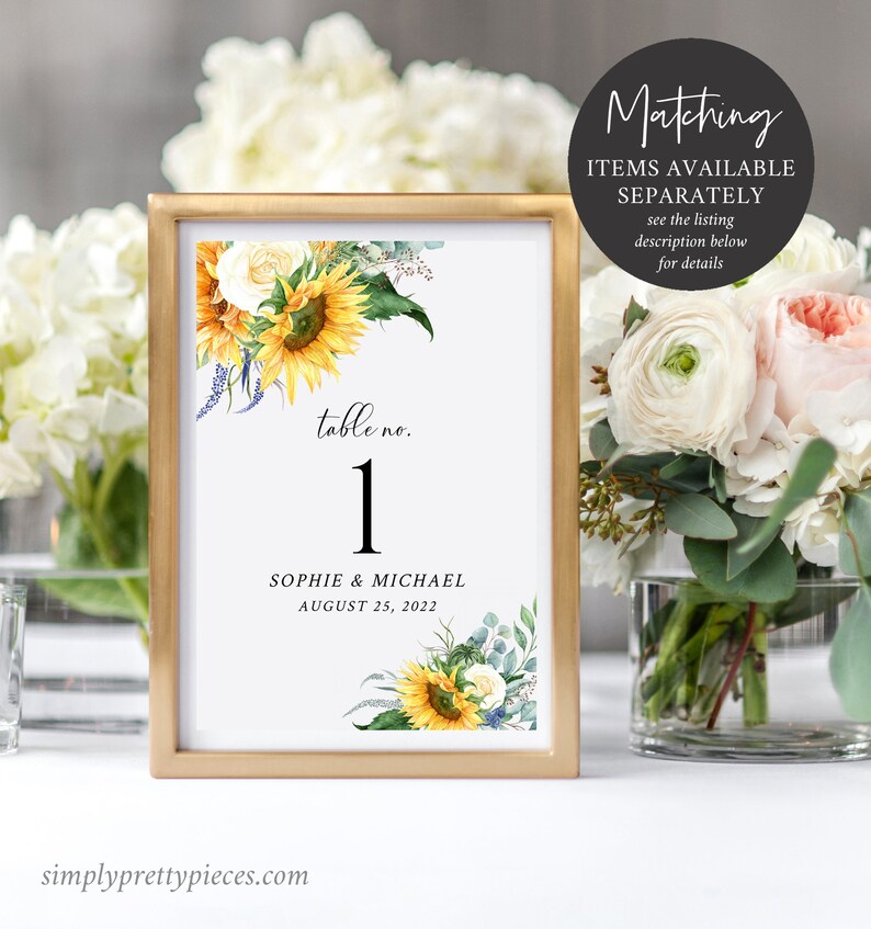 May include: A gold-framed table number card with a sunflower and white rose floral design. The card reads "table no. 1" and "Sophie & Michael August 25, 2022".
