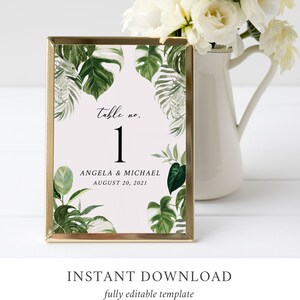 May include: A gold-framed table number card with a tropical leaf design. The card features the text "table no. 1" and the names "Angela & Michael" with the date "August 20, 2021".